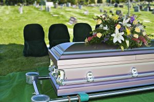 Elegant silver casket with floral arrangement at a cemetery grave site, mourners' chairs in the background.