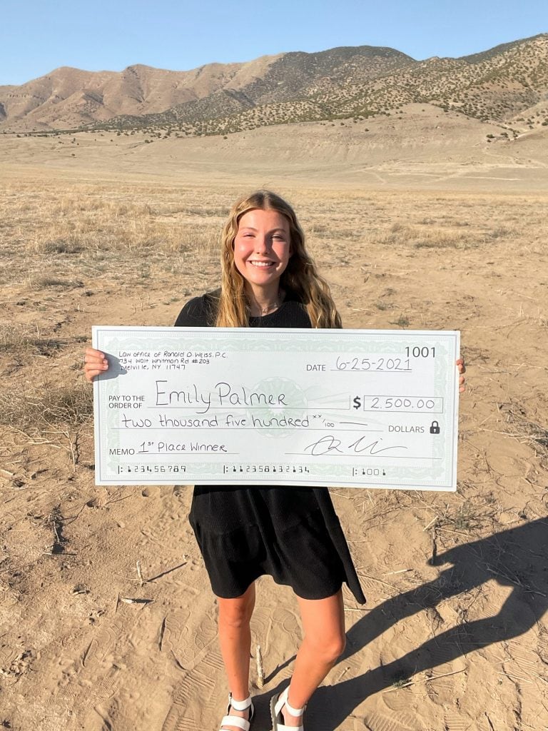 A person standing outdoors in a dry, mountainous area holding a large ceremonial check for $2,500 made out to Emily Palmer, awarded by the Law Office of Ronald D. Weiss P.C. as the 1st Place Winner on June 25, 2021