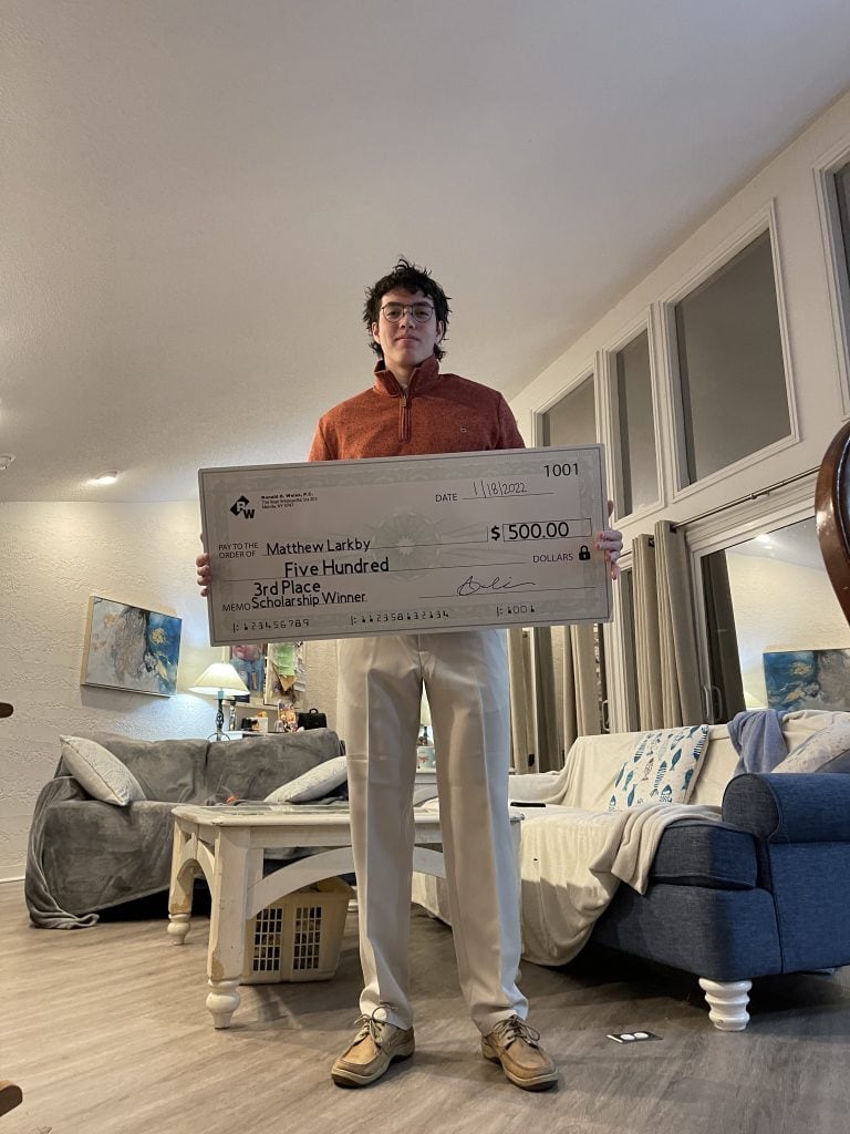 A person stands in a living room holding a large ceremonial check from the National Society of High School Scholars, awarded to Matthew Larkby for winning 3rd place in a scholarship competition worth $500.00, dated April 18, 2020.