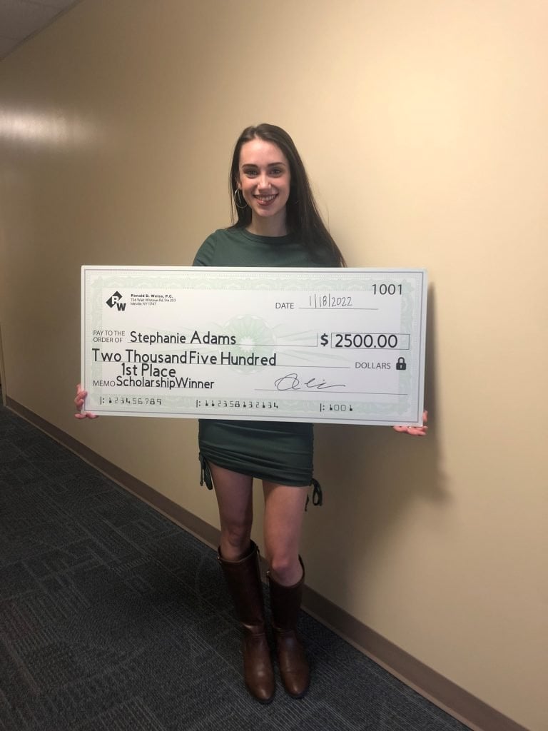 A person stands in a hallway holding a large ceremonial check made out to Stephanie Adams for $2,500.00, awarded for winning 1st place in a scholarship competition on November 8, 2022.
