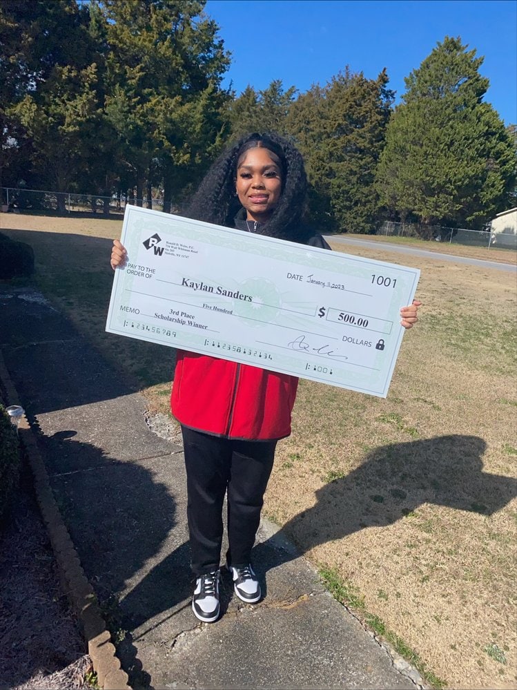 $500 scholarship check awarded to young girl holding oversized check outdoors.