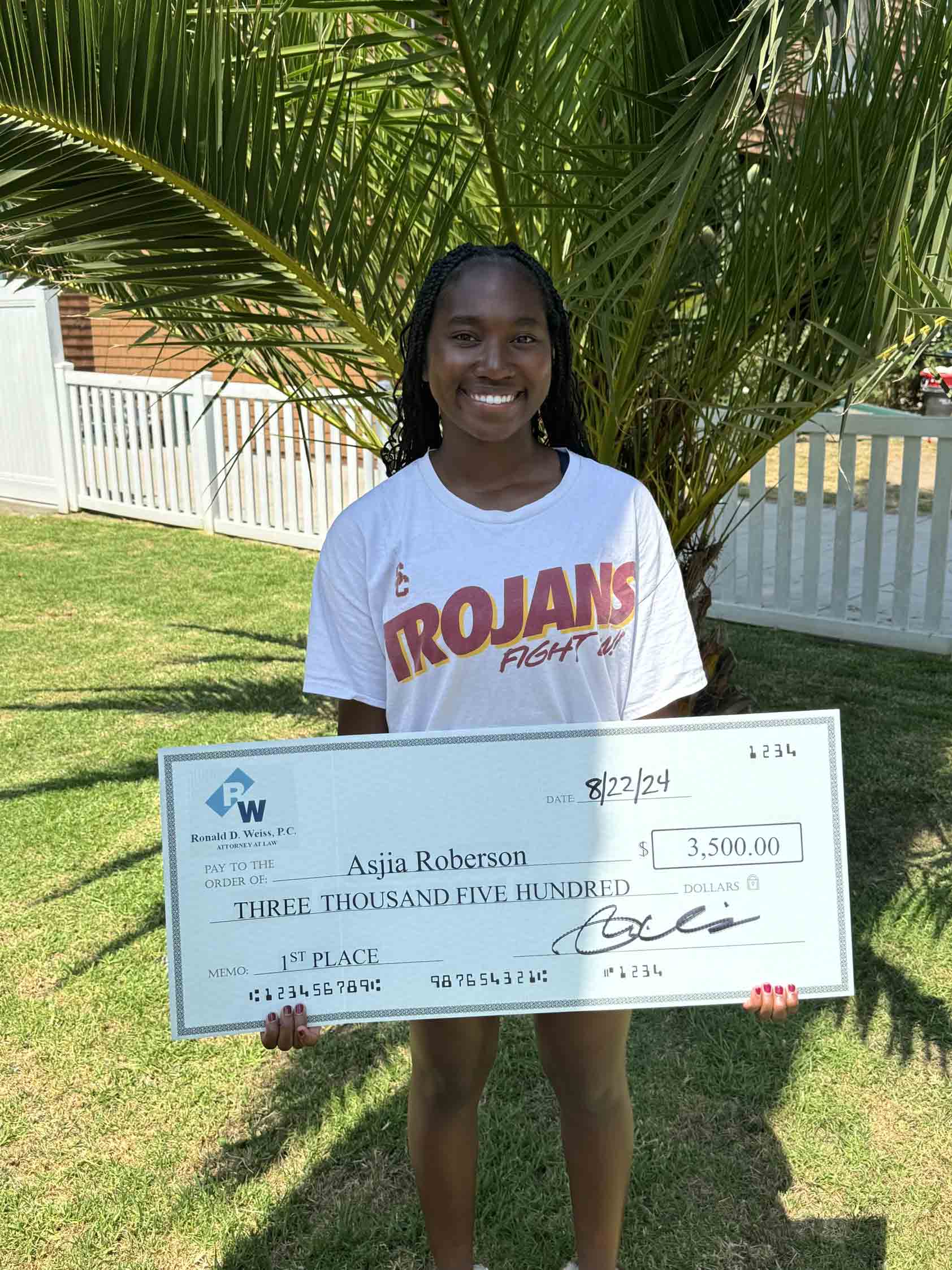 Large check held by smiling woman in front of tropical plant, celebrating financial achievement or award.
